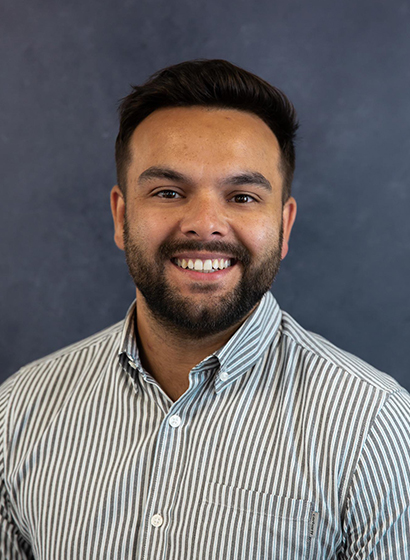 Portrait of Luke Savva, wearing a blue and white striped shirt against a dark mottled navy backdrop