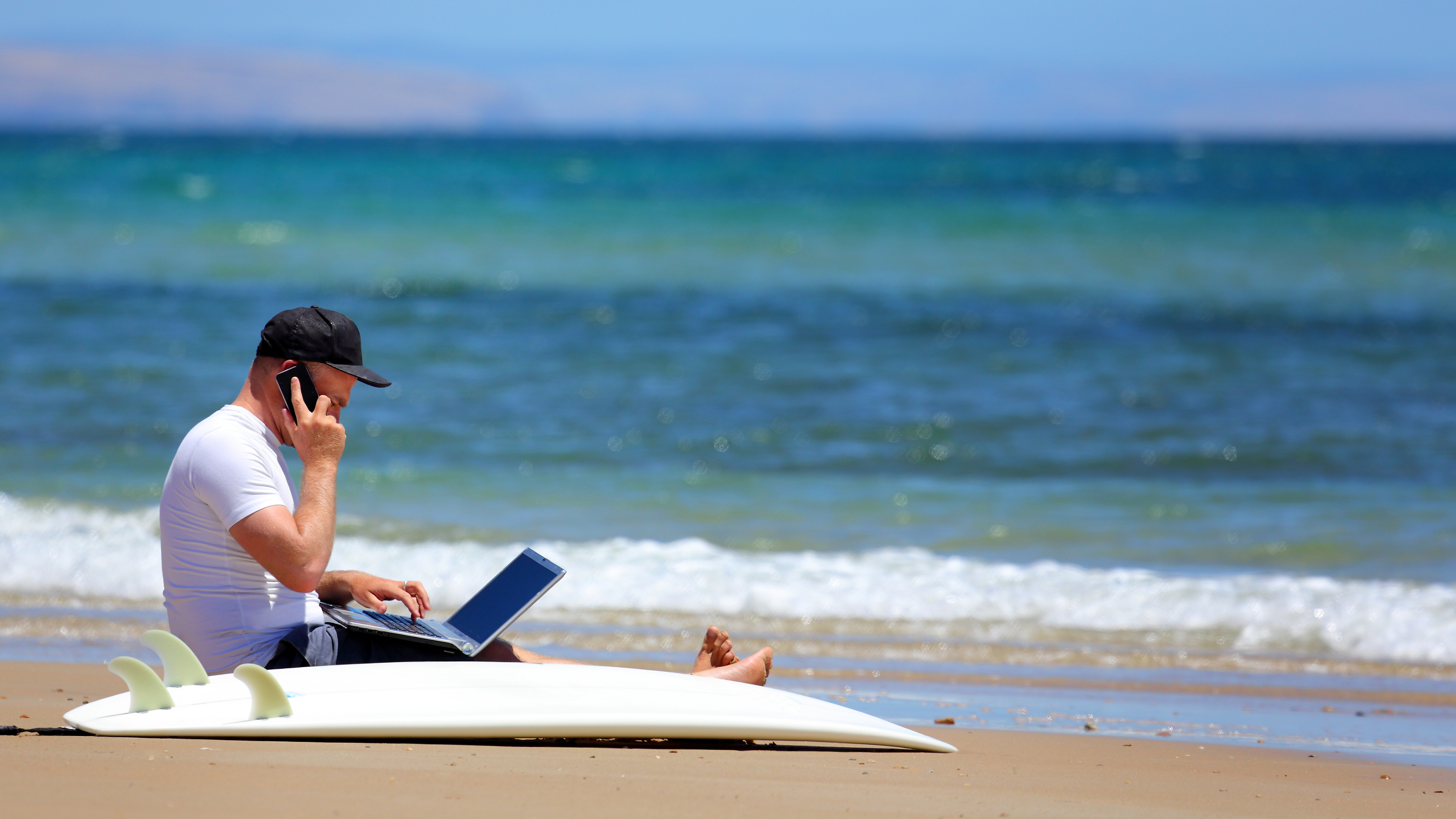 surfer in cap sitting on Australian beach, talking on smartphone with laptop in front of him