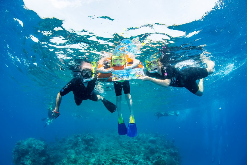 Three people snorkeling underwater above a coral reef. A child in the middle wearing an orange float and yellow snorkel holds a colorful sign reading ‘Great Barrier Reef’. On either side, two adults wearing black wetsuits and snorkel gear swim alongside, smiling toward the camera