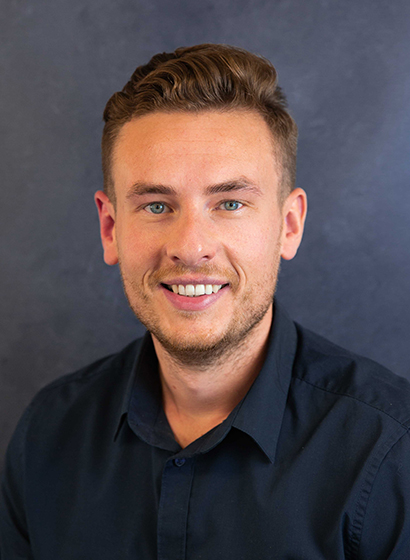 Portrait of Tom Kerwin, a blond haired tanned man in a navy shirt against a dark mottled navy backdrop