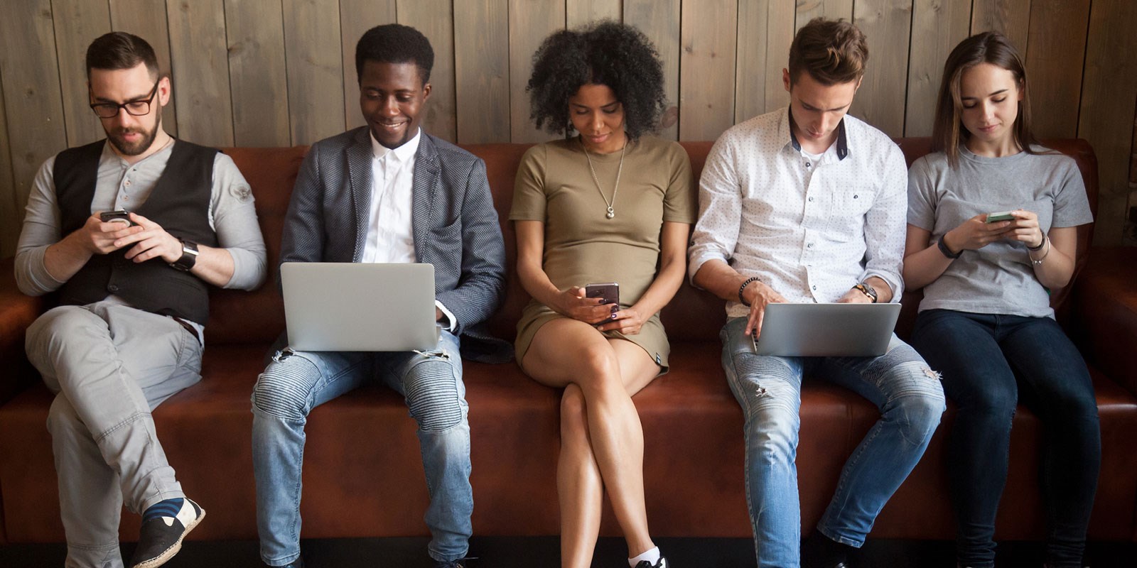 5 young people sitting in a row, all looking at laptops or smartphones